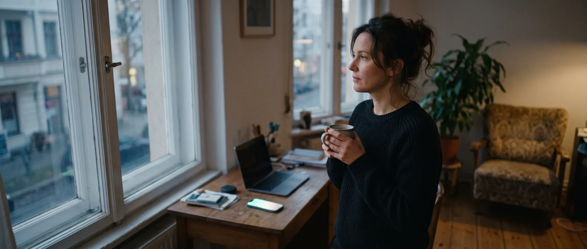 A business owner standing away from her desk at dusk with a coffee, while her phone keeps working behind her